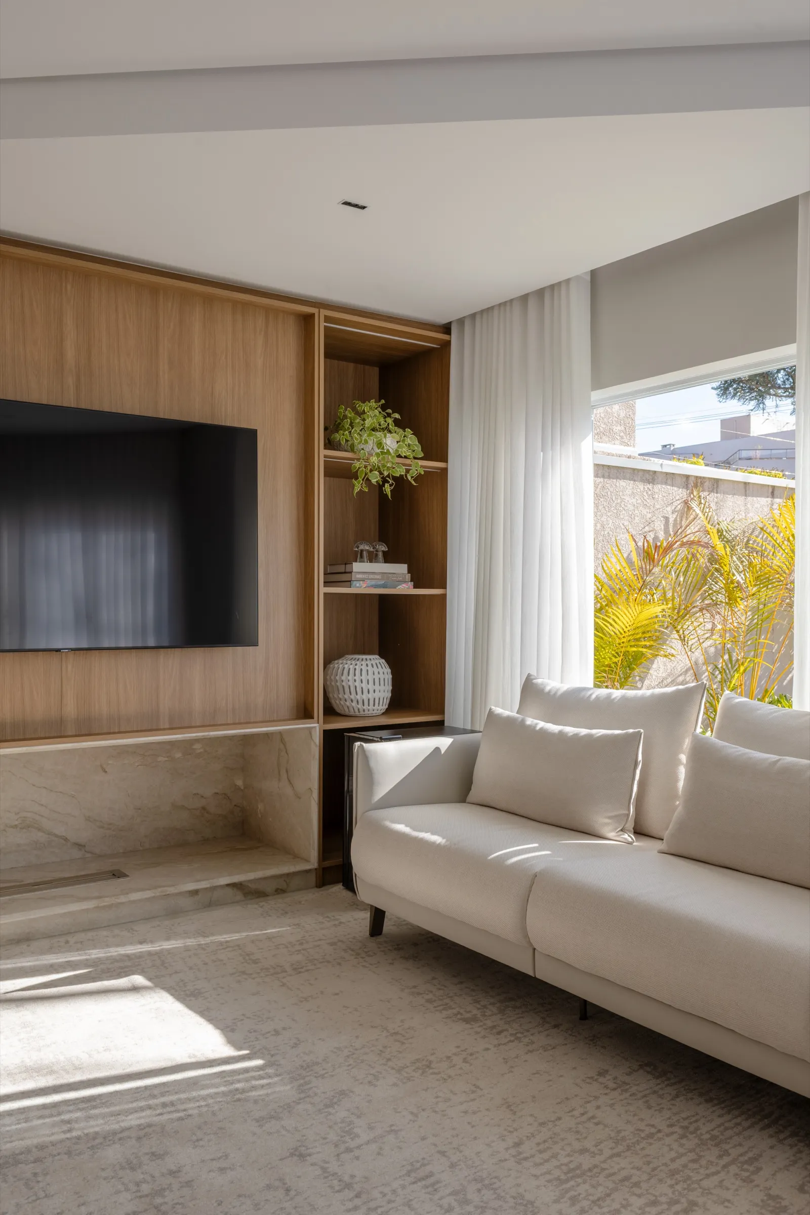 Sofá claro diante de painel de madeira com TV e janela com jardim