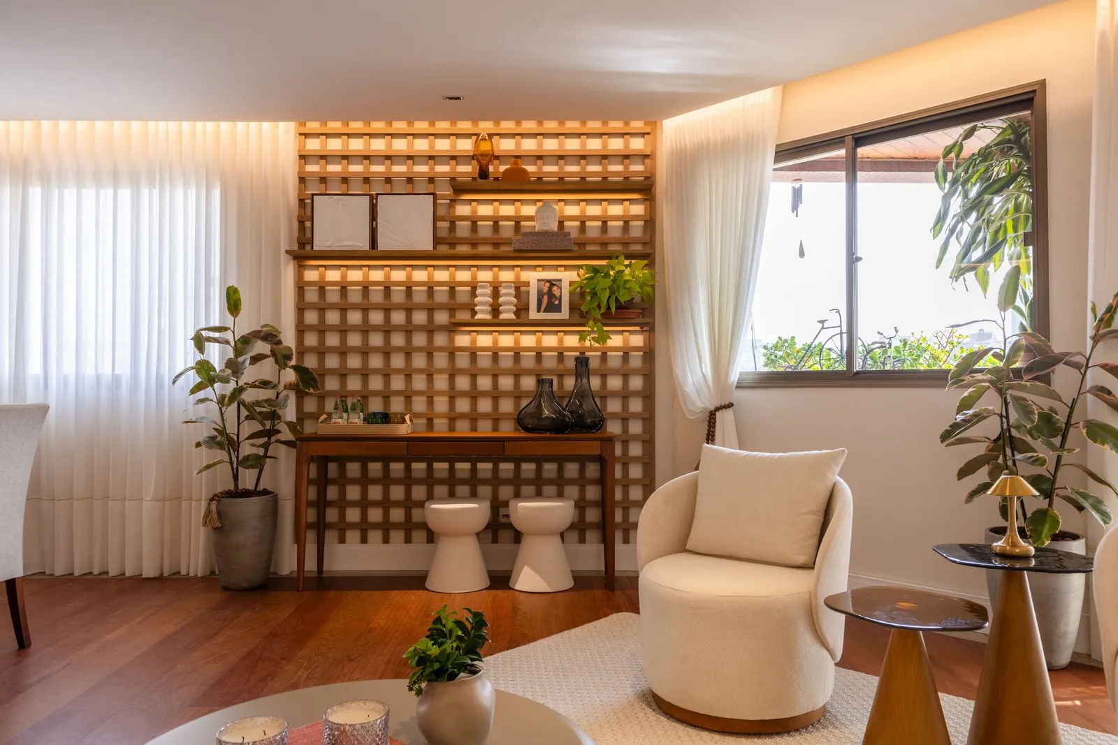 Sala do Apartamento Grécia com estante de madeira iluminada, poltrona clara e janela