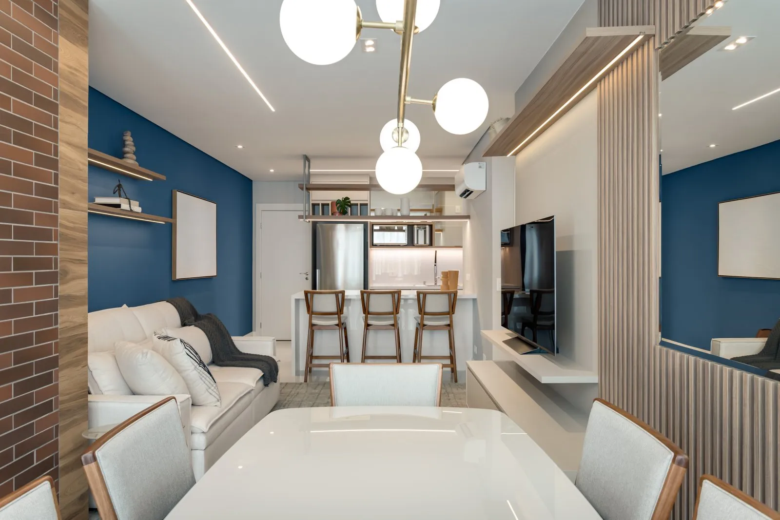 Sala integrada com parede azul, tijolinho, pendente de globos e mesa branca