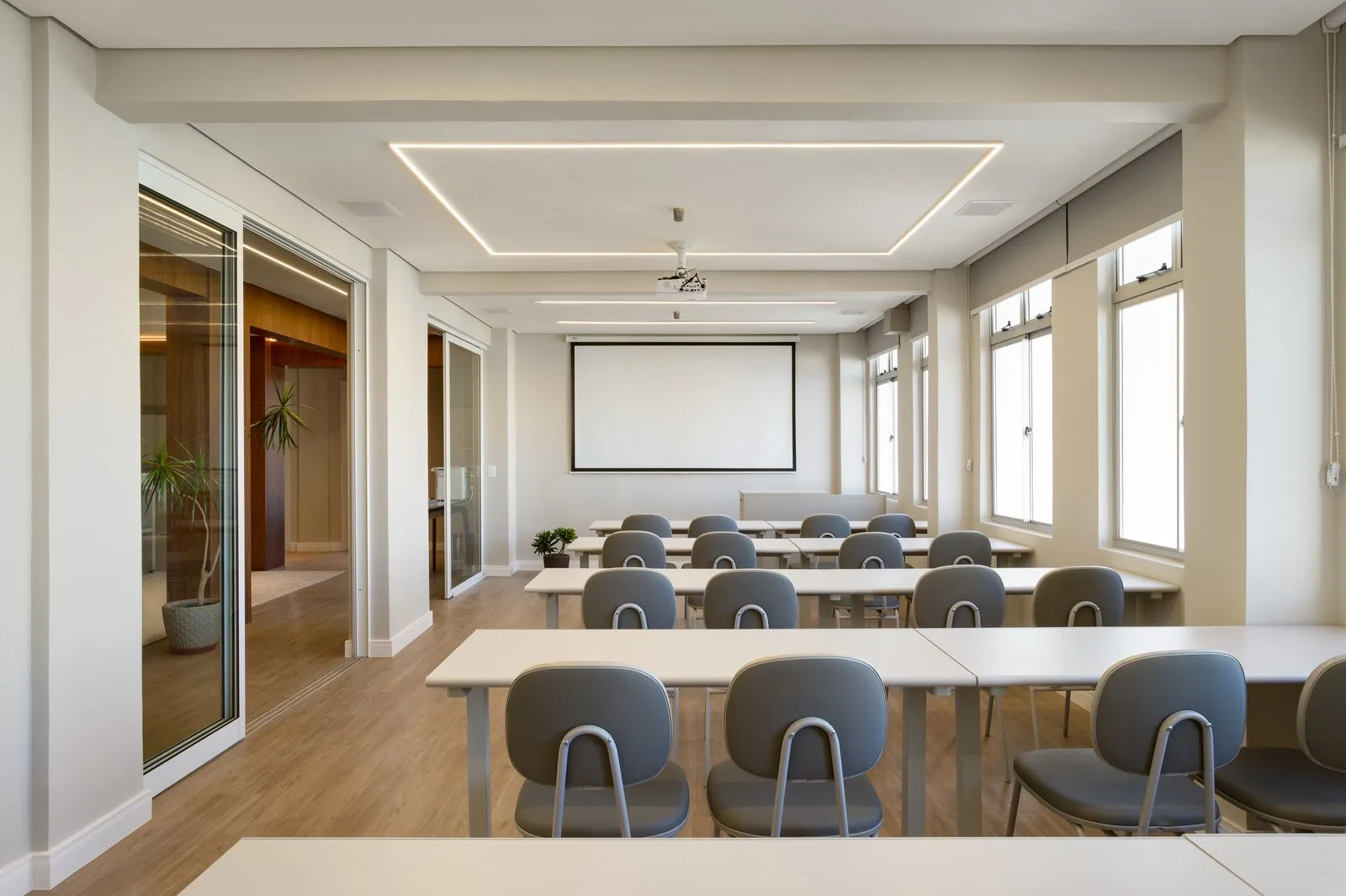 Sala de aula com mesas brancas, cadeiras cinza e tela de projeção