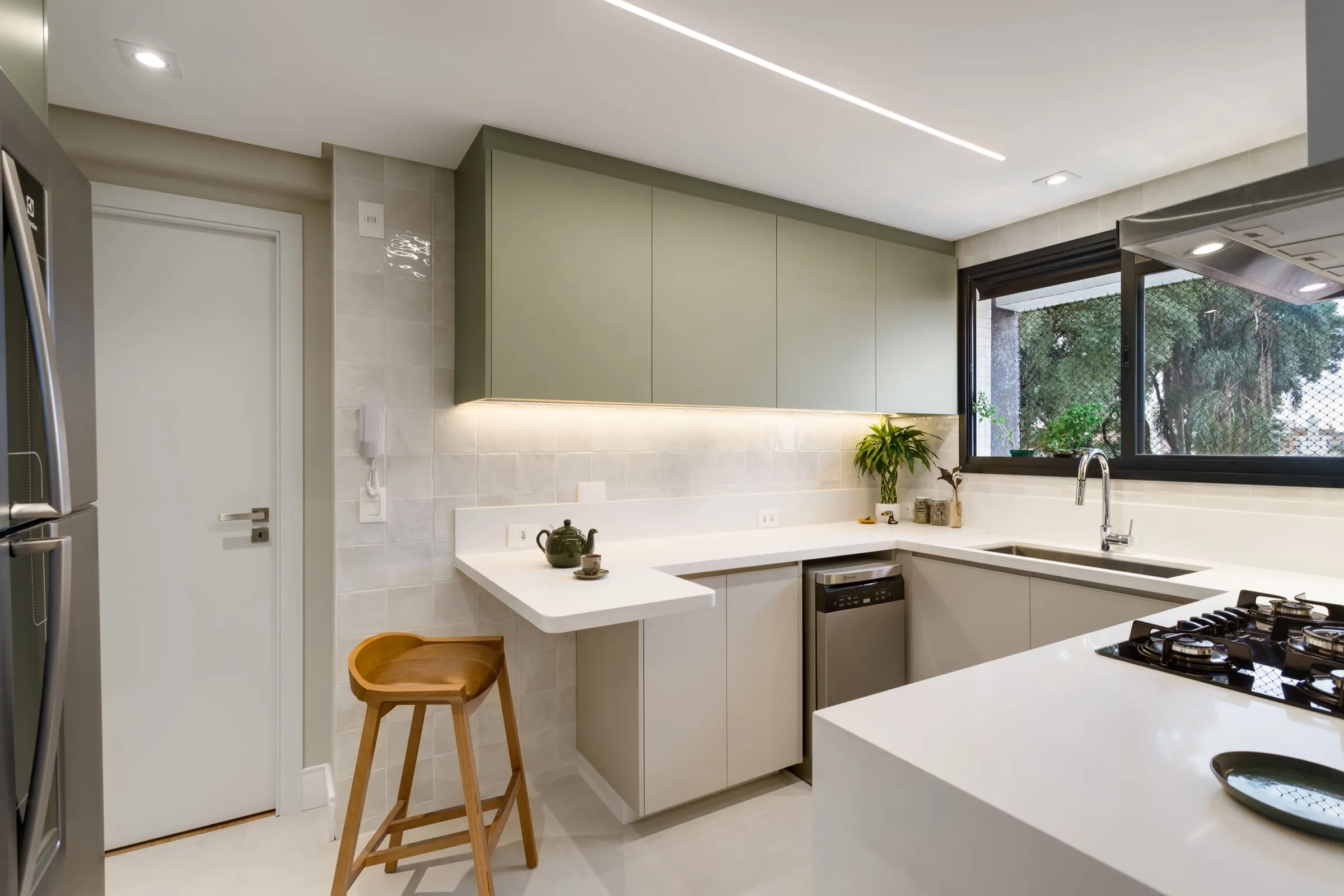 Cozinha em U com armários verdes, bancada branca e banqueta de madeira