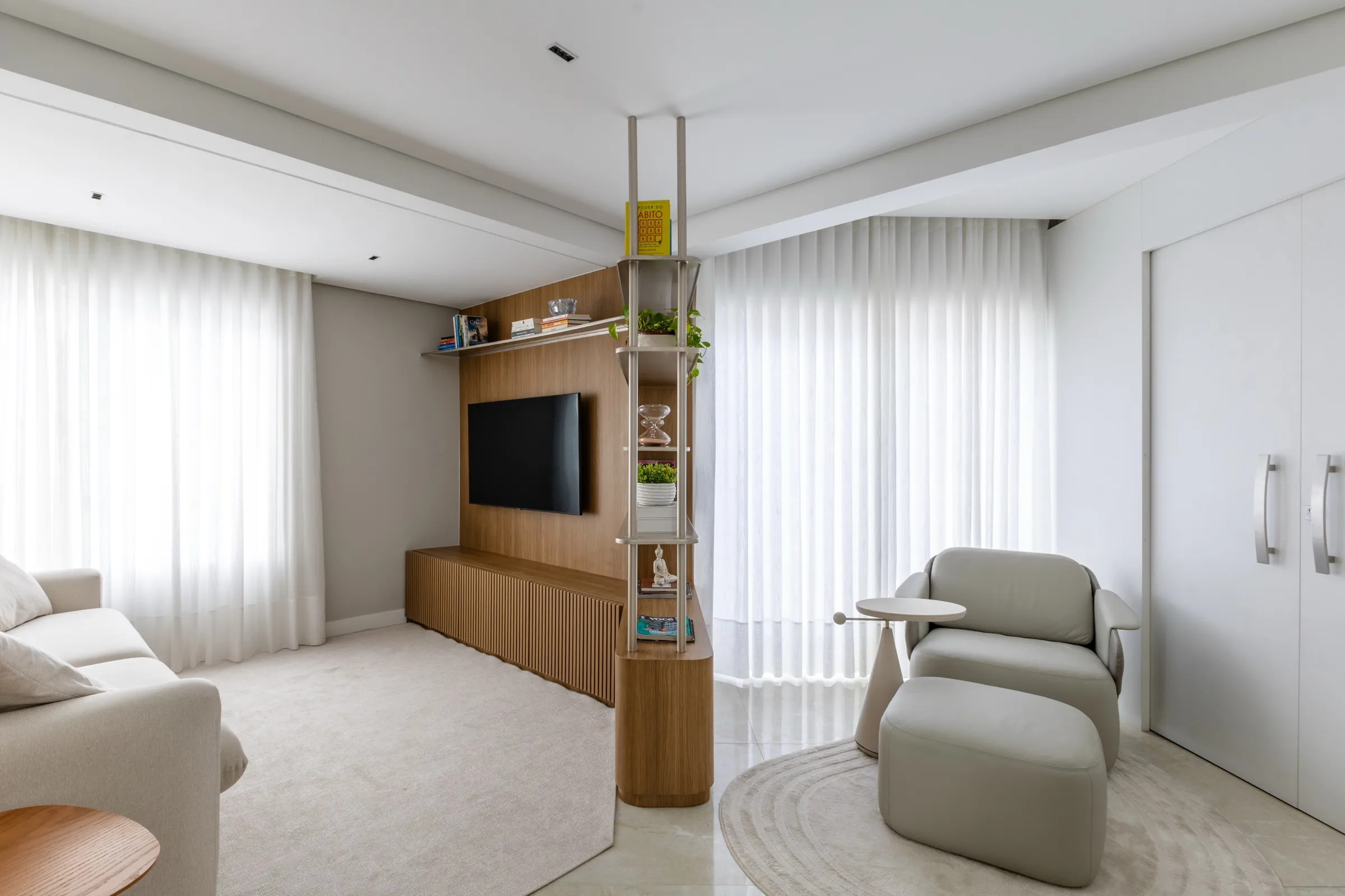 Sala com rack ripado, estante vazada e poltrona com pufe diante da cortina