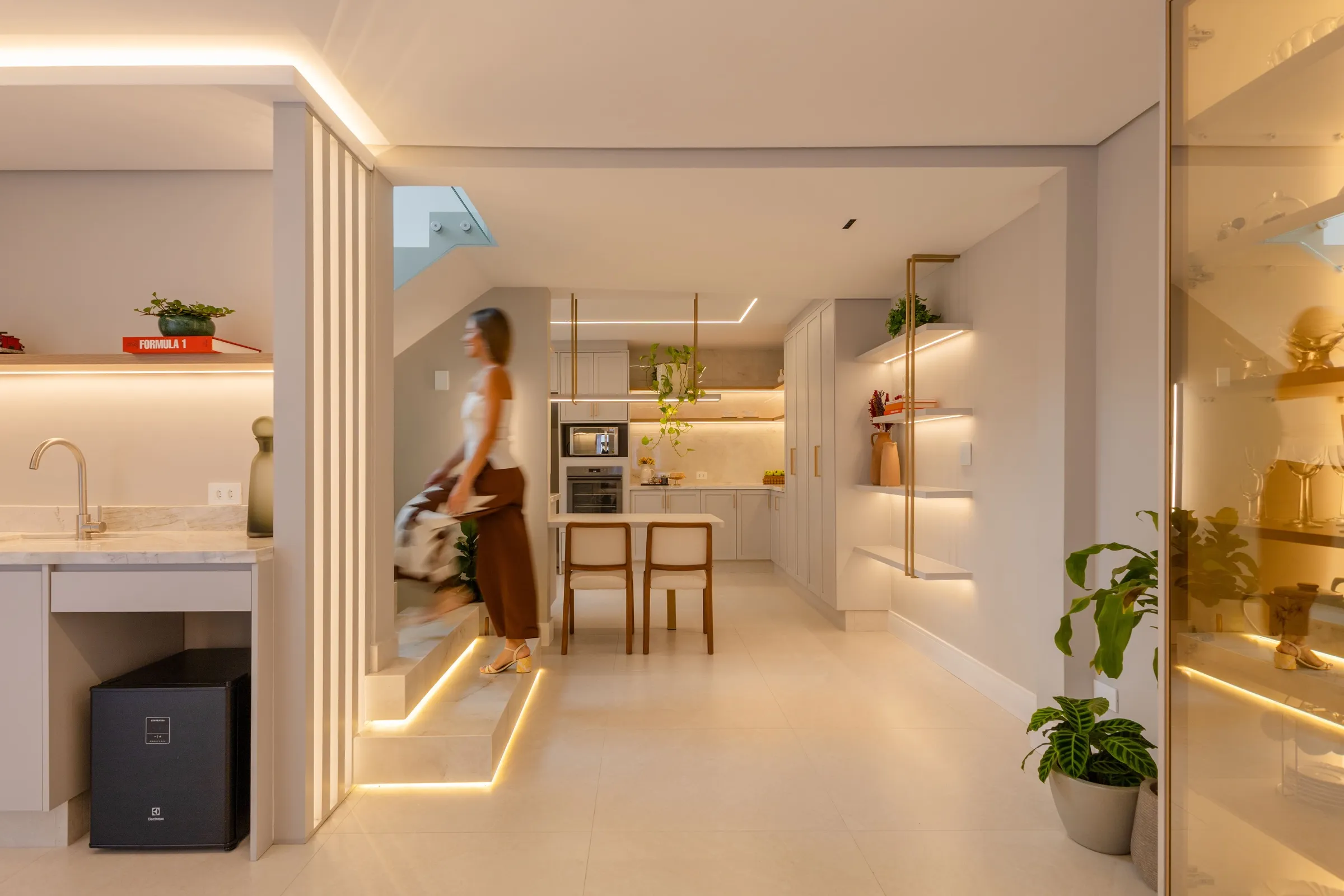 Ambiente com escada iluminada, cozinha ao fundo e prateleiras de vidro