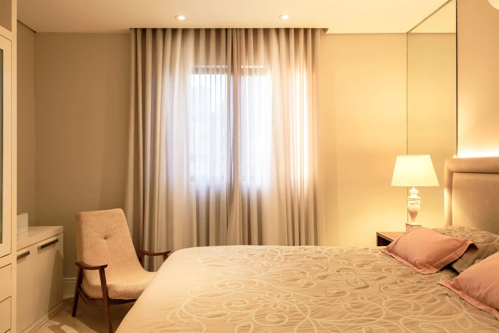 Quarto com cama de cabeceira estofada, poltrona e cortina diante da janela