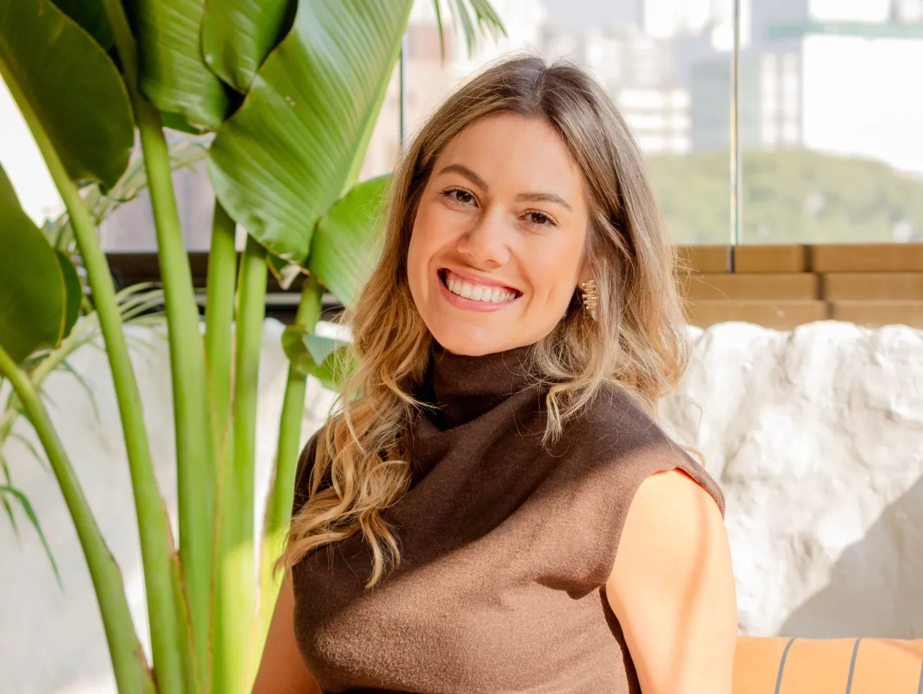 Rafaela Bender, arquiteta, sorrindo ao lado de uma planta, de blusa marrom
