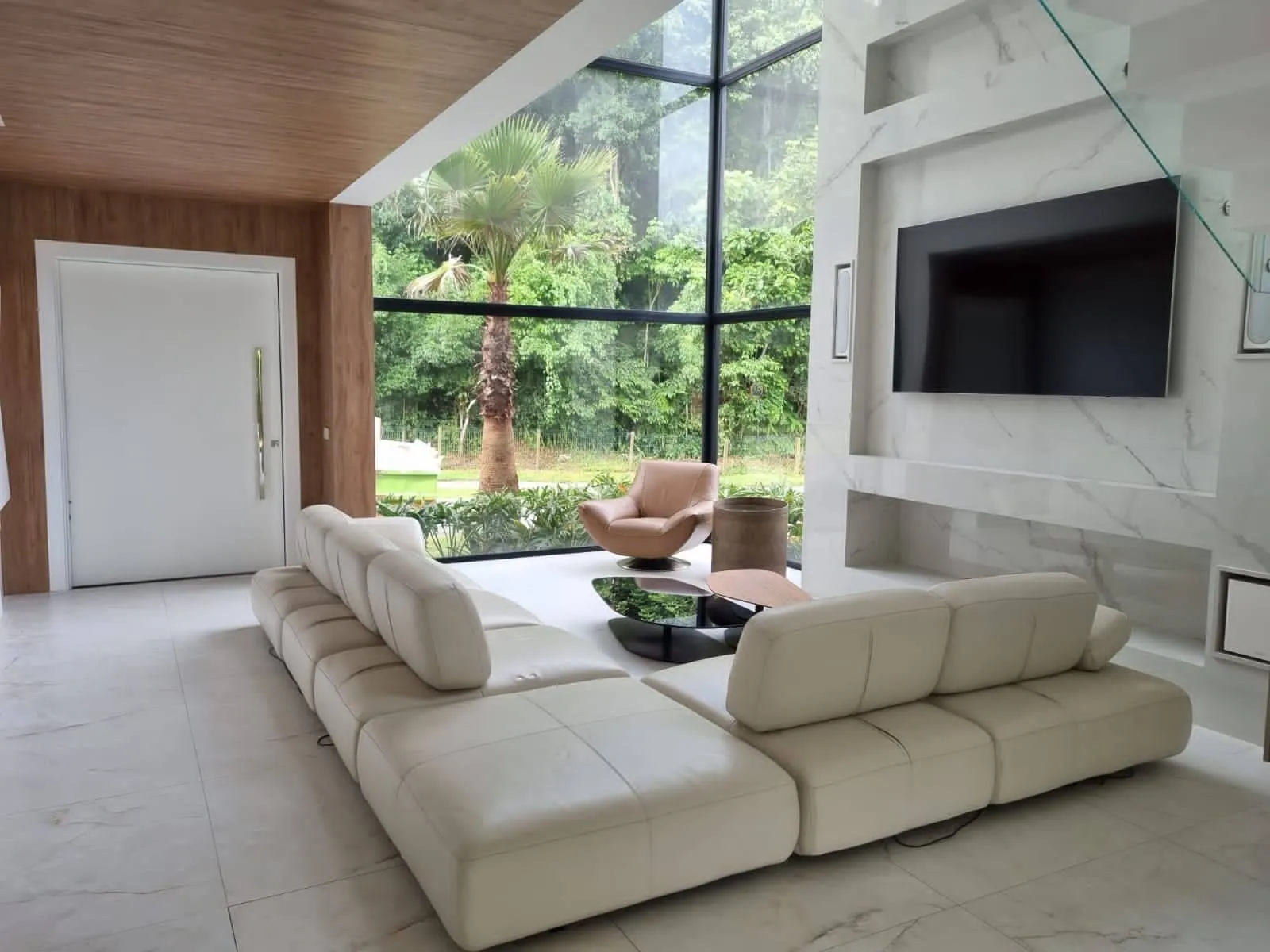 Sala de estar com sofá branco em L, painel de TV em mármore e janela de vidro do chão ao teto voltada para o jardim